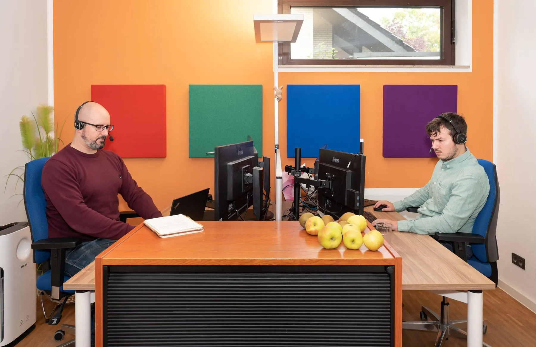 Zwei Entwickler mit Headsets an ihren Arbeitsplätzen, auf dem Tisch frisches Obst