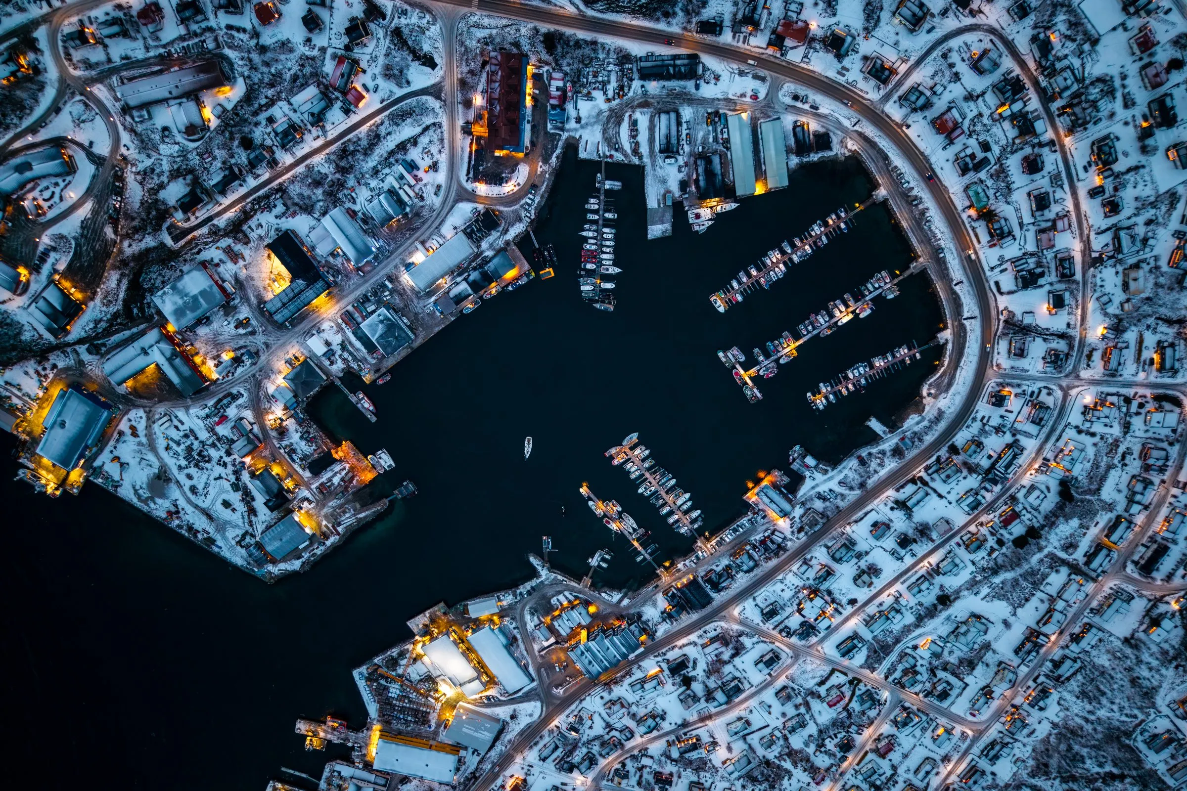 Luftbild senkrecht von oben auf einen verschneiten Hafen bei Nacht, beleuchtete Stege und Hallen rund um das dunkle Hafenbecken