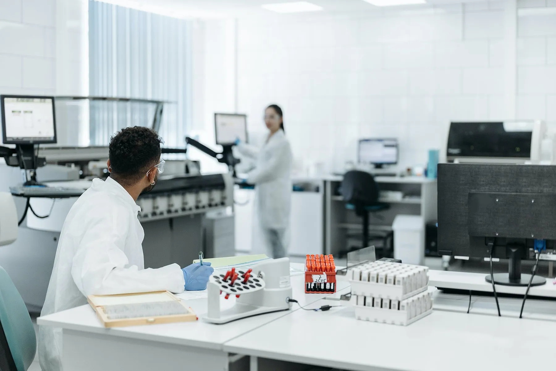 Labor mit Analysegeräten und Bildschirmarbeitsplätzen, im Vordergrund ein Mitarbeiter mit Probenröhrchen
